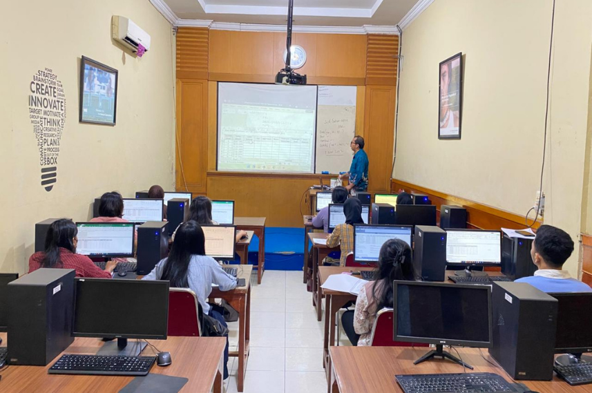 Instructor at the front of a computer lab giving a presentation to students at desks with desktop PCs and a projected screen behind him.