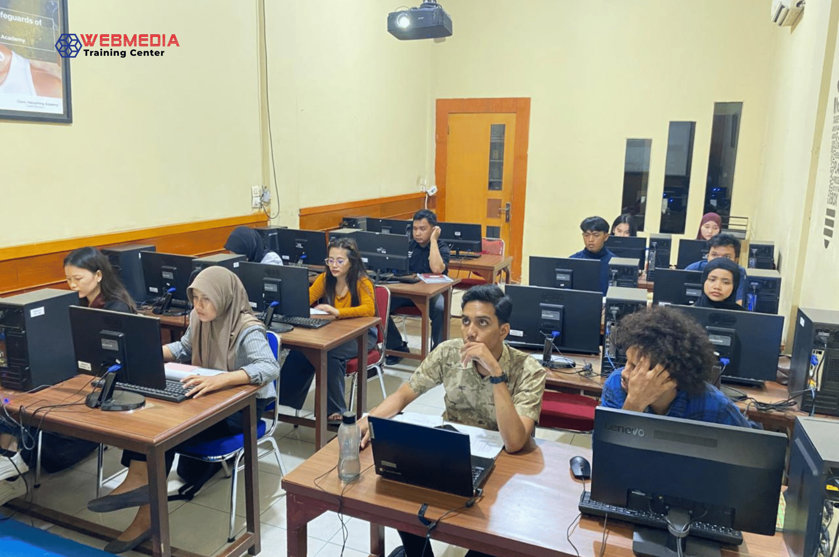 Students in a computer lab working at desks with desktop PCs; WEBMEDIA Training Center logo visible in the corner.
